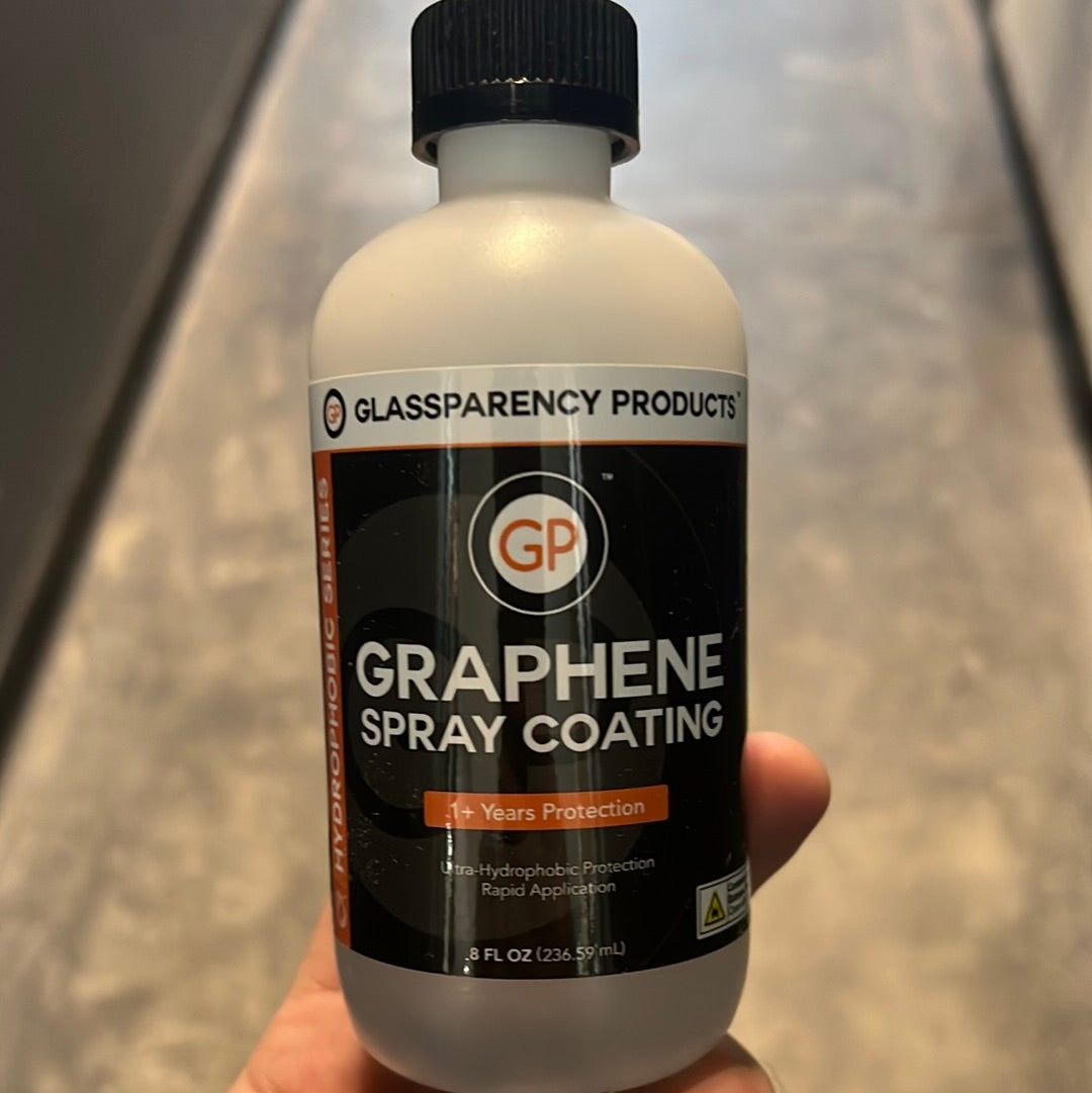 A hand holds GP Graphene Spray coating bottle with black label, featuring a close-up of the logo and a yellow triangle on the label.