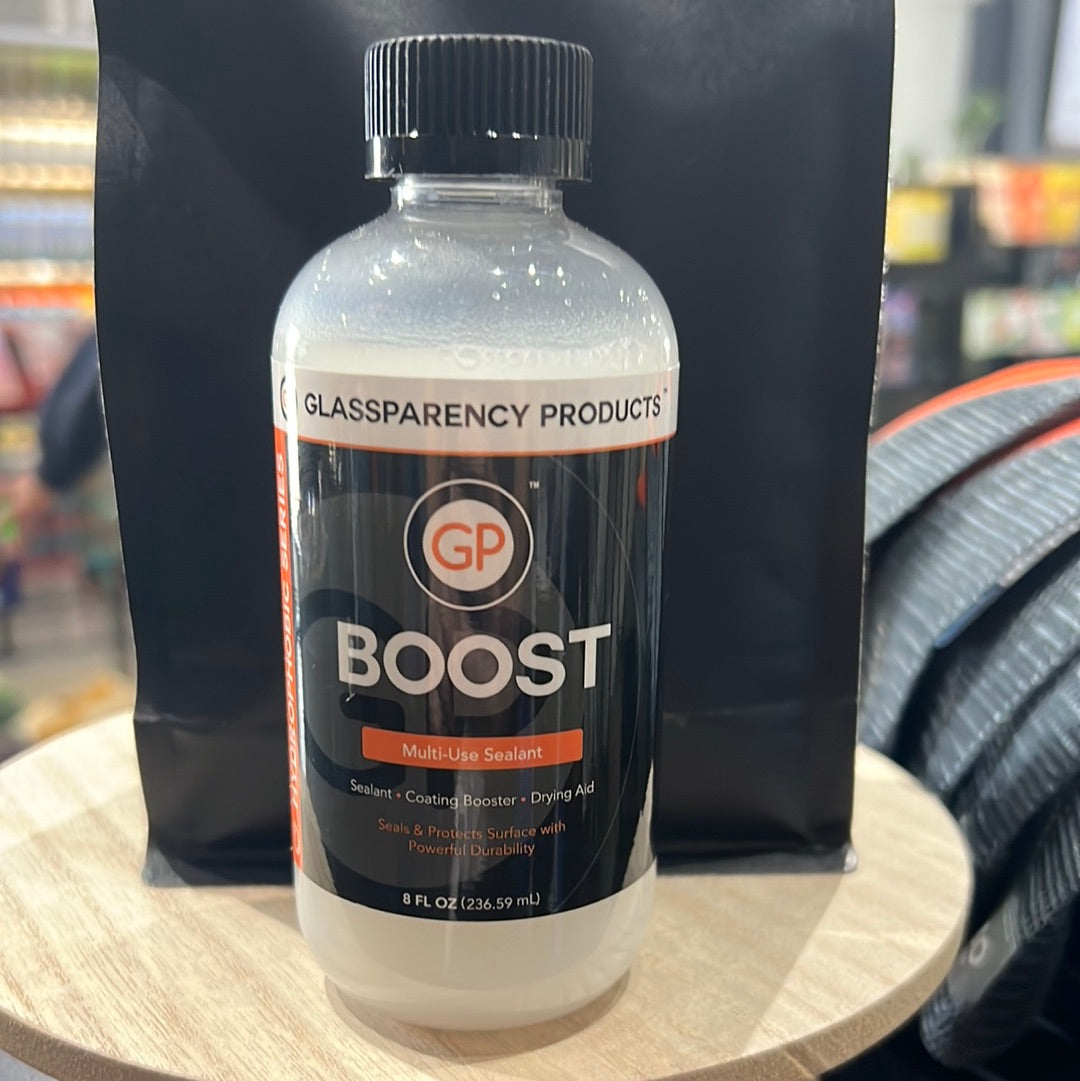 A close-up of GP Boost, a bottle of white liquid solution for ceramic coated cars, displayed on a table with a blurred hand in the background.