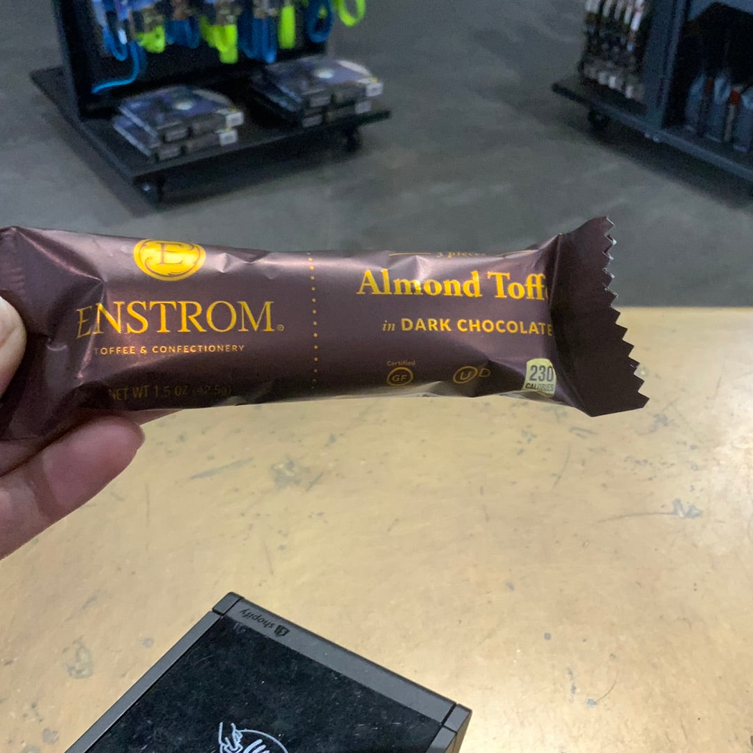 A hand holding Enstrom almond toffee, a chocolate bar. Package of almonds visible. Close-up of device in background. Snack and candy.