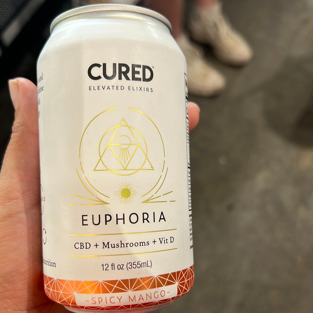 A hand holds a Cured Spicy Mango can with white and gold design, featuring a symbol and label on a close-up aluminum can.