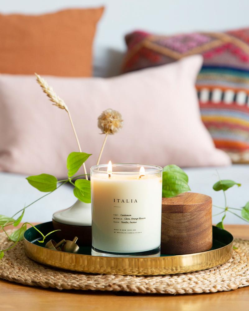 A candle named Italia Candle on a tray, surrounded by lit candles, a wooden bowl, and plants. Fragrant blend of cardamom, clove, vanilla, incense, and orange blossom. Made in Brooklyn with soy wax.