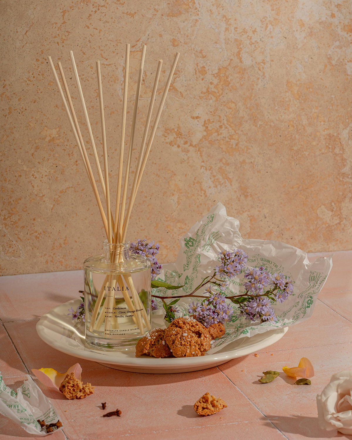 A plate with sticks and flowers, a glass jar, cookies, and a rose, embodying the fragrant Italia Reed Diffuser. Includes wooden stopper and 8 rattan reeds.