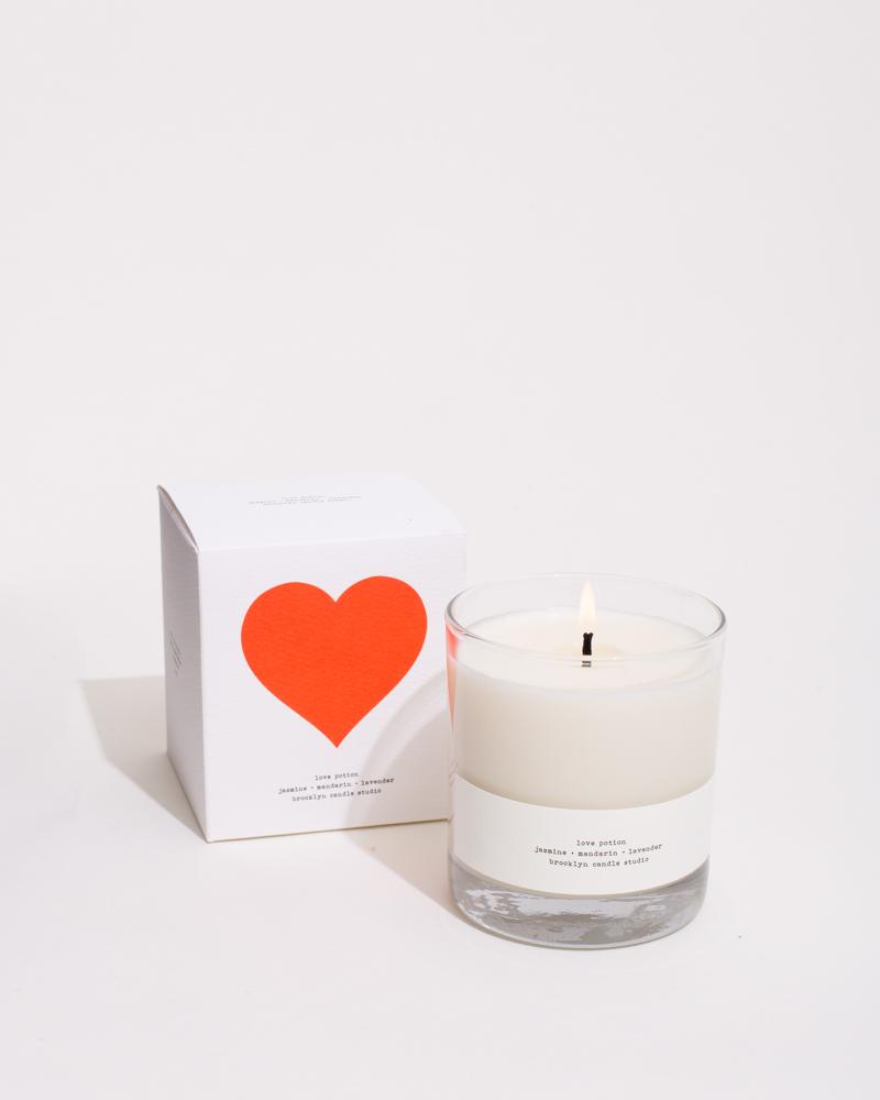 A candle named Love Potion in a glass tumbler with a textured label, next to a white box with a red heart. Handcrafted in Brooklyn with jasmine, lavender, and mandarin scents.