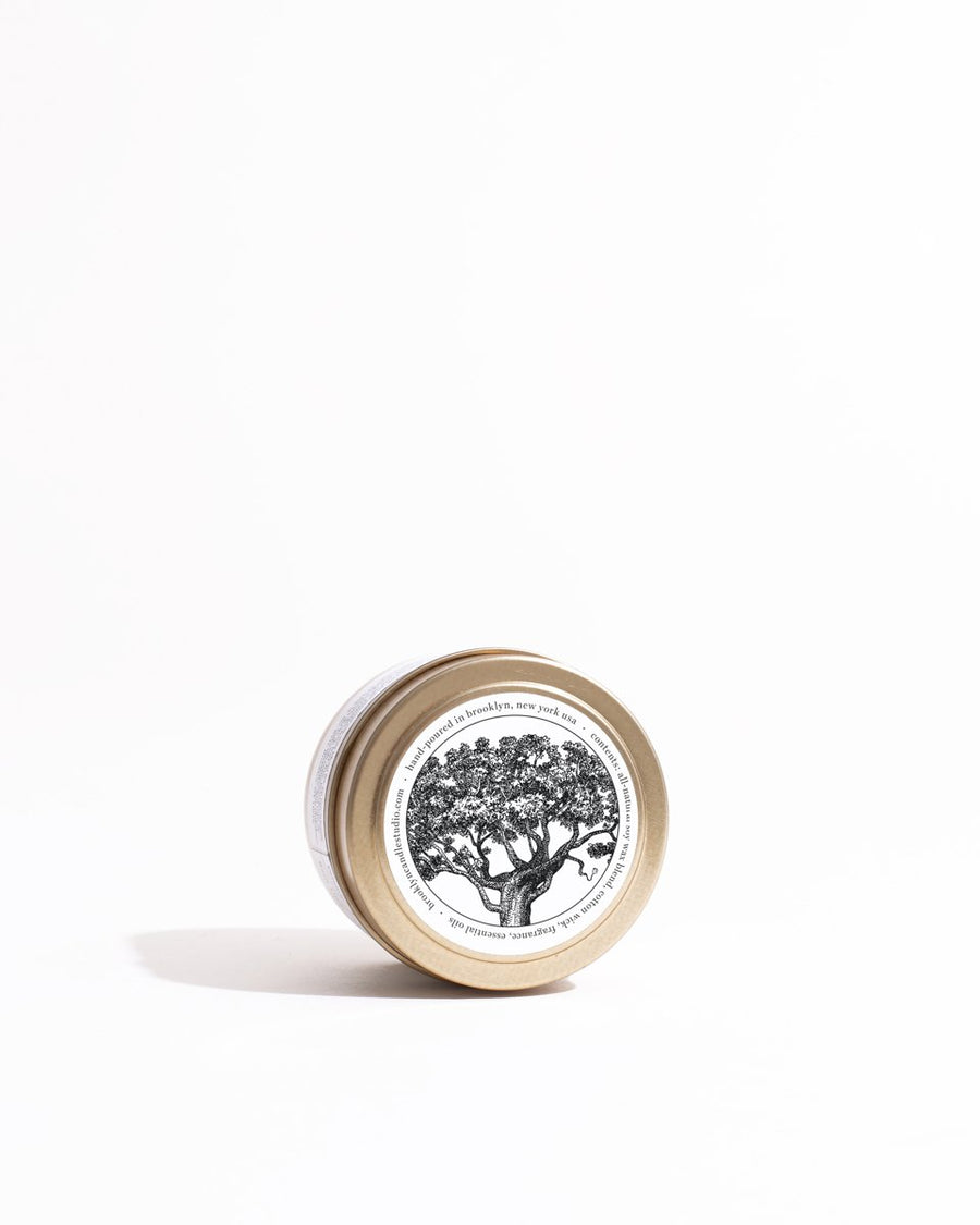 A round gold travel candle tin with a tree drawing label, embodying the Palo Santo scent. Eco-friendly soy wax, essential oils, and cruelty-free.